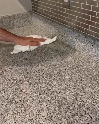 Hand wiping a light gray speckled granite countertop with a white cloth beside a tile backsplash