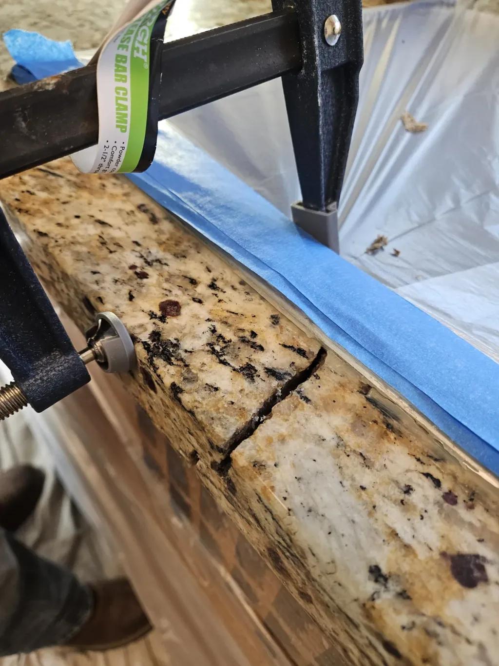 Crack in a dark granite countertop edge held closed with clamps, the surrounding stone masked with blue tape
