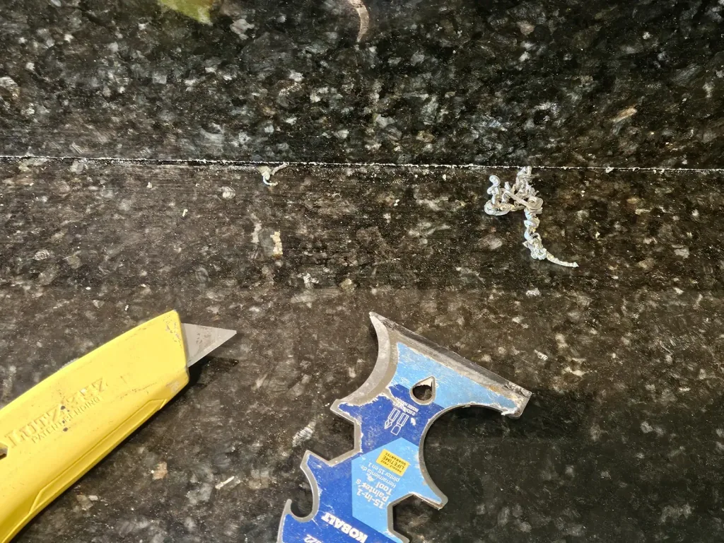 Scraper lifting old caulk and failed filler from a dark granite countertop