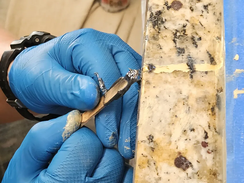 Gloved hands pressing tinted repair resin into a break along the edge of a light speckled granite countertop