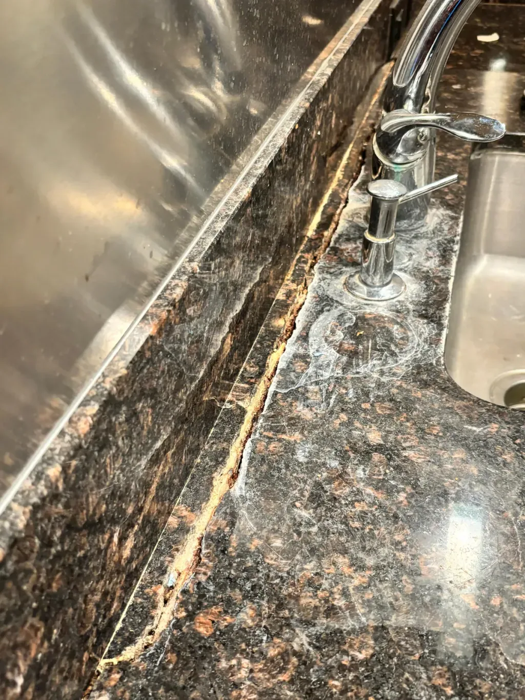Open crack running along a dark granite sink rail behind the faucet