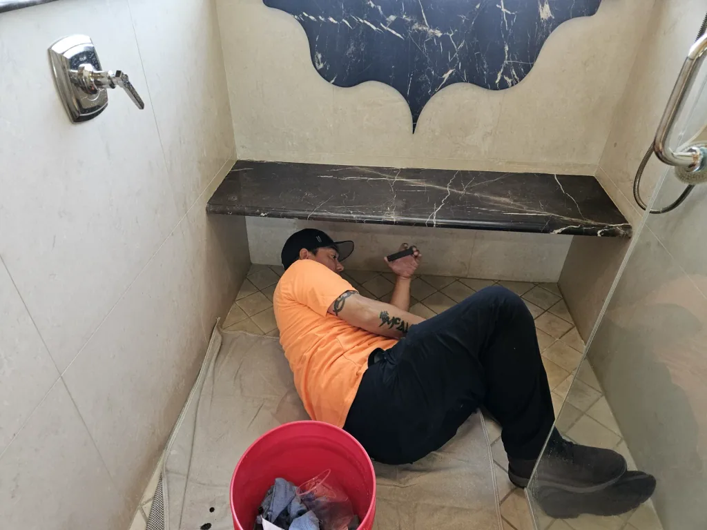 Stain Wranglers technician working at floor level inside a marble shower beside a dark stone bench
