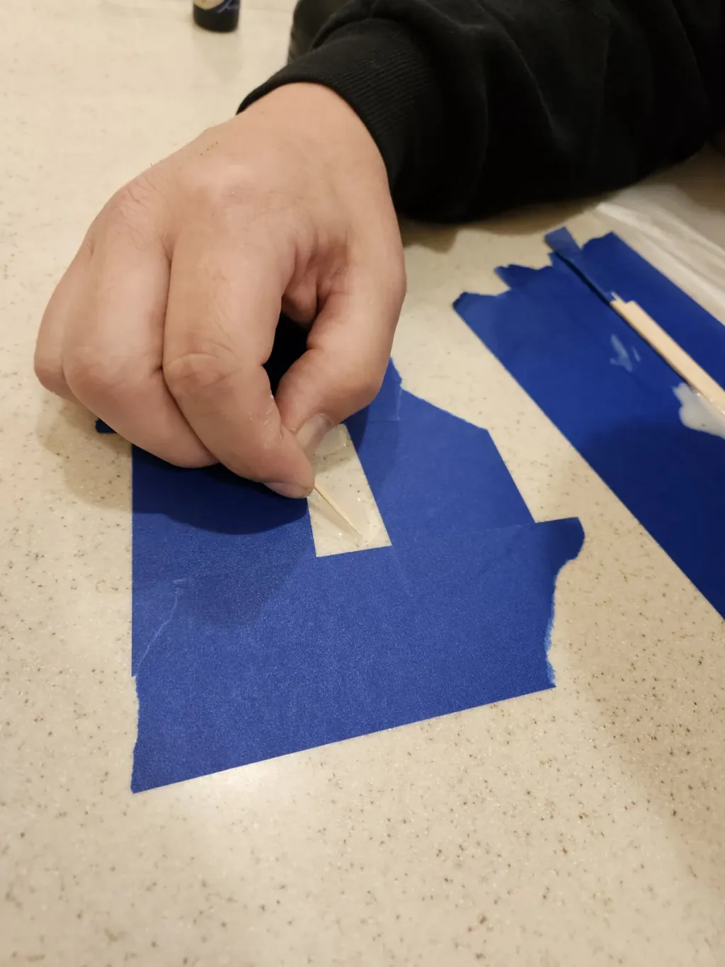 Hand pressing tinted resin into a chip through a window cut in blue masking tape on a speckled quartz countertop