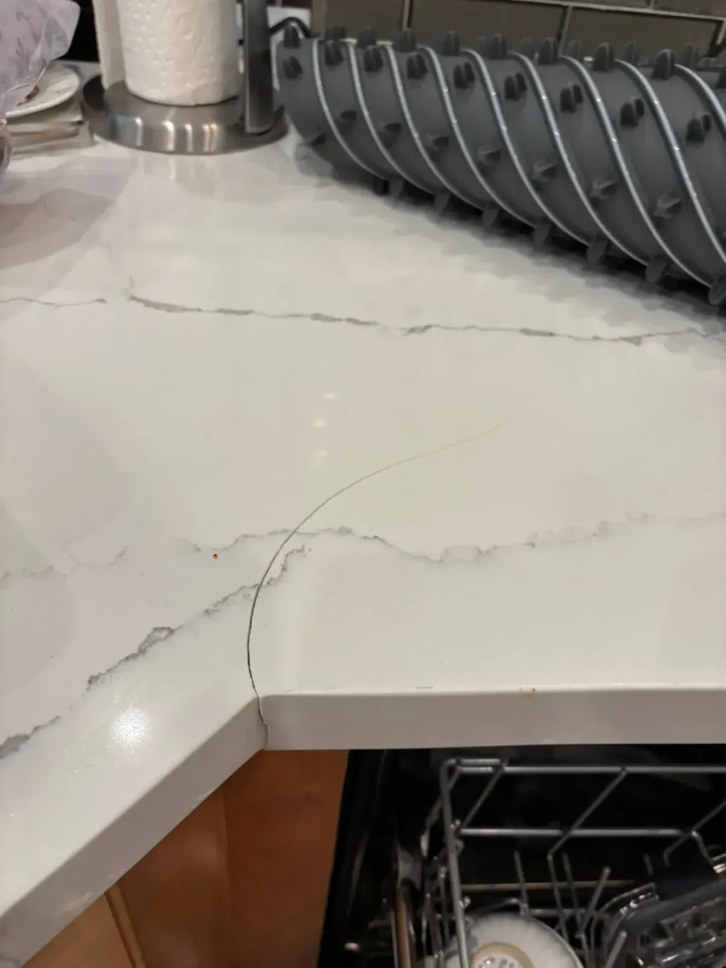 Hairline crack running across the corner of an L-shaped white quartz countertop above an open dishwasher