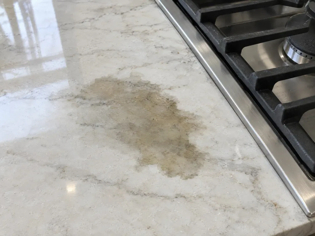 Dark, soft-edged oil stain in a pale quartzite countertop beside a cooktop