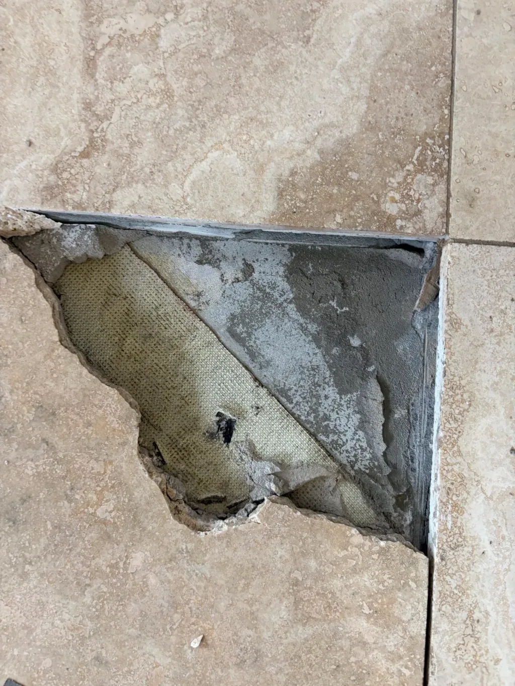 Cracked travertine tile partly broken out, showing the thinset bed and a mesh membrane underneath