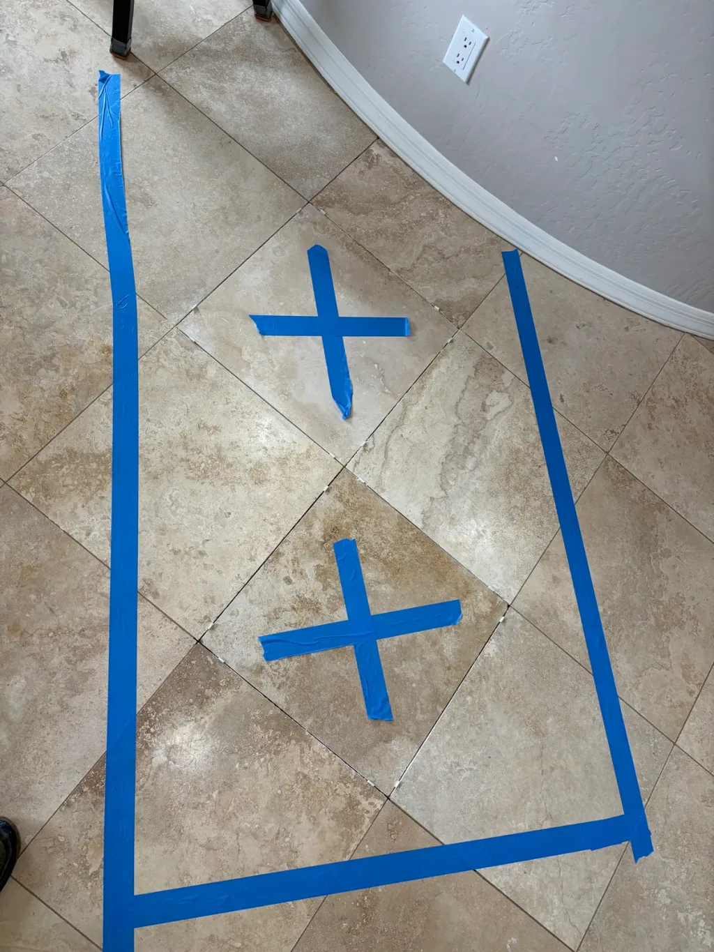 Blue tape marking two cracked travertine tiles for replacement on a diagonal floor