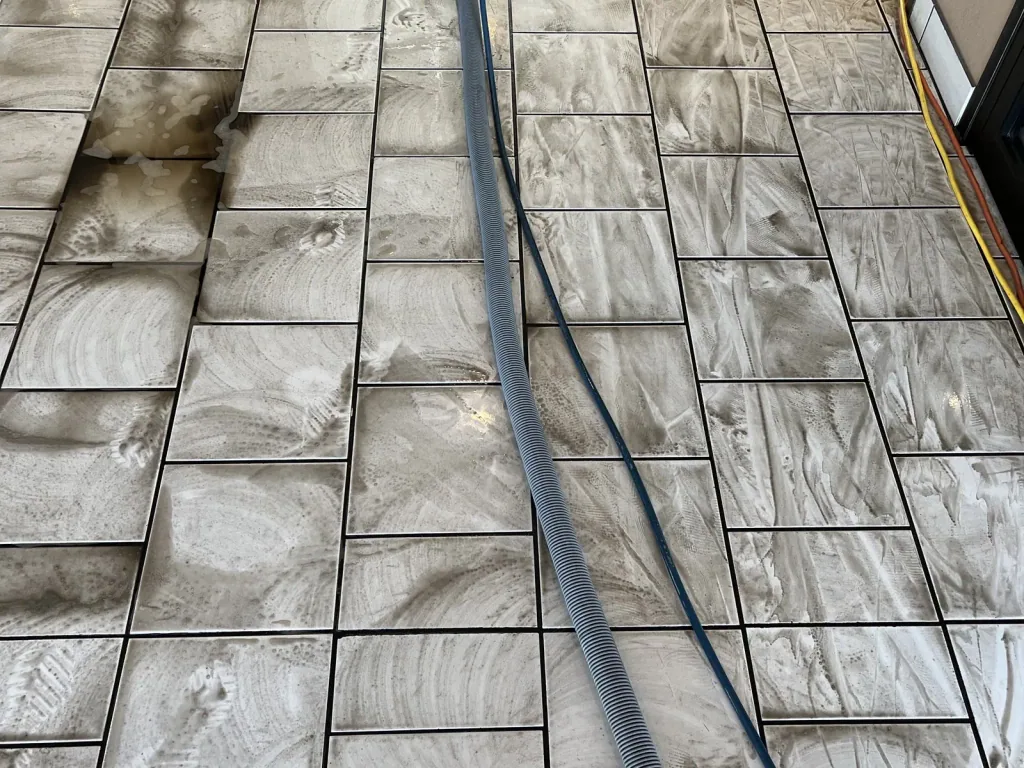 Stone tile floor partway through cleaning, with clean tiles beside tiles still dark with soil and the extraction hose running across them